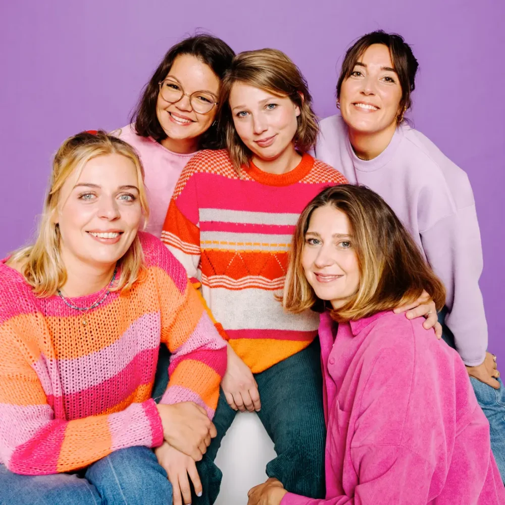 Gruppenfoto von fünf Frauen in bunt gestreiften Pullovern vor lila Hintergrund
