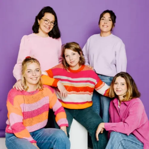 Fünf lächelnde Frauen in bunten Pullovern vor lila Hintergrund