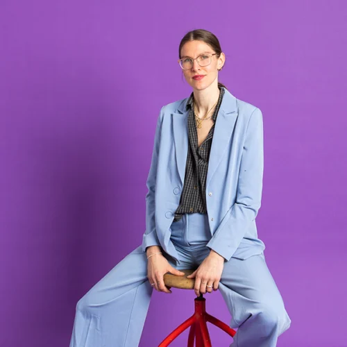 Frau in blauem Anzug vor lila Hintergrund sitzt auf Hocker