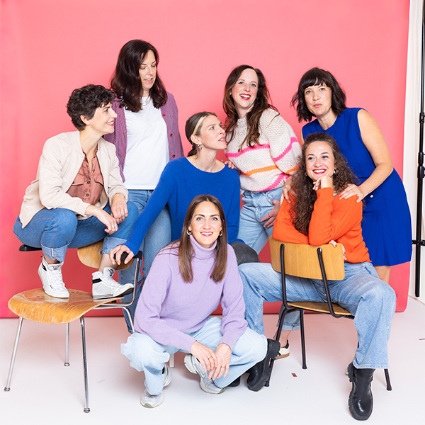 Sieben Frauen posieren lächelnd vor rotem Hintergrund auf Stühlen