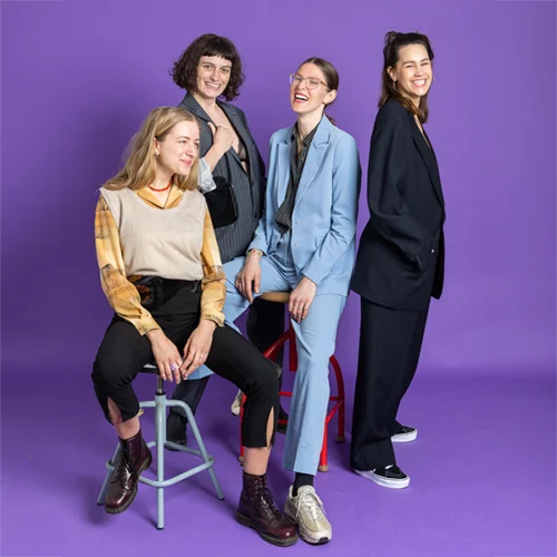 Vier stilvolle Frauen posieren lächelnd vor lila Hintergrund