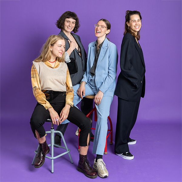 Vier stilvolle Frauen posieren lächelnd vor lila Hintergrund