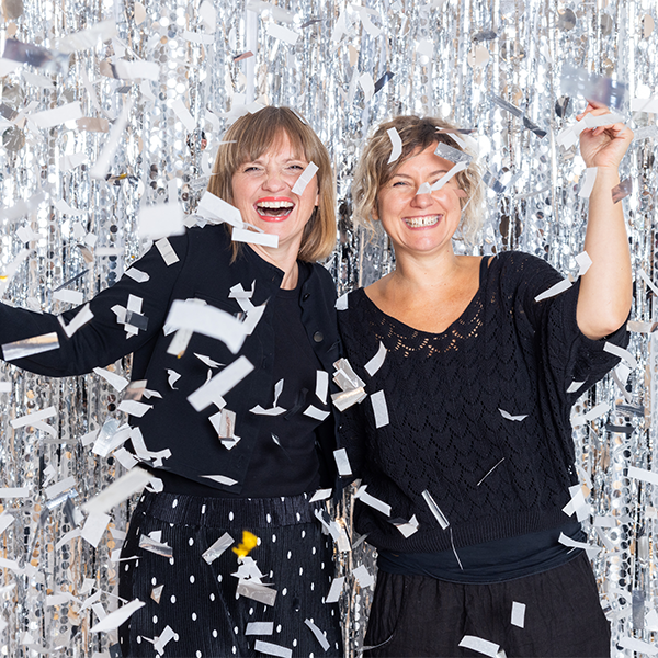 Zwei lachende Frauen feiern mit Konfetti vor glitzerndem Hintergrund