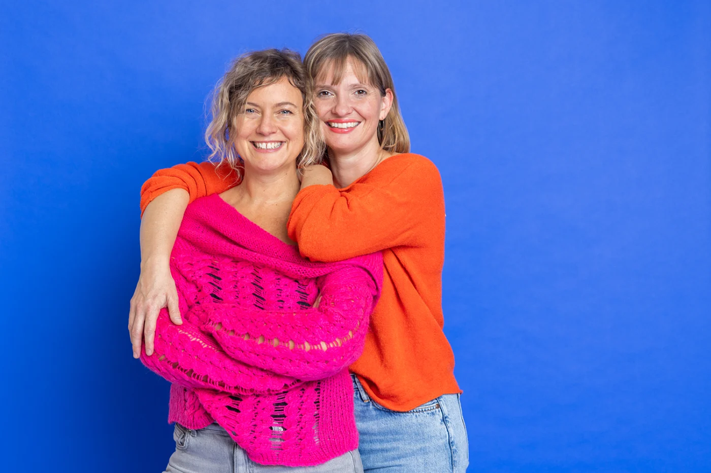 Zwei lächelnde Frauen in farbenfroher Kleidung vor blauem Hintergrund